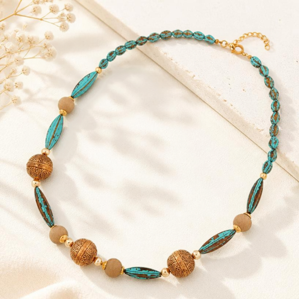 Türkis-patina-farbene Perlenkette mit altgoldfarbenen Musterperlen und beigebraunen Rundperlen auf hellem Untergrund mit zarten weißen Blüten.