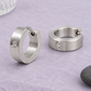 Matte Edelstahl-Klappcreolen mit klaren Glassteinen, eine auf dem Rand stehend und eine flach liegend, auf lavendelfarbenem Untergrund mit Glas und dunklem Kiesel.
