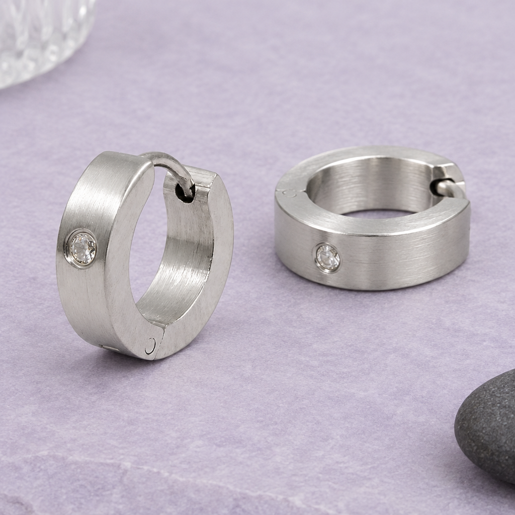 Matte Edelstahl-Klappcreolen mit klaren Glassteinen, eine auf dem Rand stehend und eine flach liegend, auf lavendelfarbenem Untergrund mit Glas und dunklem Kiesel.