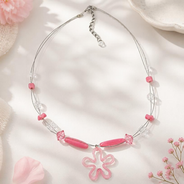 Rosa Drahtkette mit transparenten Perlen und offenem Blütenmotiv auf hellem Untergrund mit rosa Blüten und heller Keramik.
