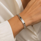 KI-generierte Frau trägt ein silberfarbenes Edelstahl-Schildband-Armband mit blanker Gravurplatte am Handgelenk zu einem weißen Oberteil.