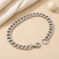 Silberfarbenes Flachpanzerarmband mit sichtbarem Karabiner und Endring auf hellbeigem Untergrund mit cremefarbenem Stoff und elfenbeinfarbener Dekokugel.