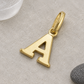 Goldener Buchstaben-Anhänger A diagonal auf warmgrauem Steinuntergrund mit hellem Glas und dunklem Kiesel.