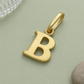 Goldener Buchstaben-Anhänger B diagonal auf salbeigrünem Untergrund mit strukturiertem Glas und warmgrauem Stein