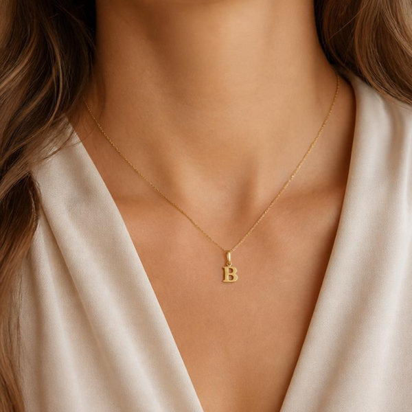 KI-generierte Frau trägt einen goldfarbenen B-Anhänger an einer feinen Kette zu einer weißen Bluse.