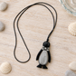 Pinguin-Pulloverkette auf hellem Holz mit Muscheln, hellen Kieseln und einer Glasschale