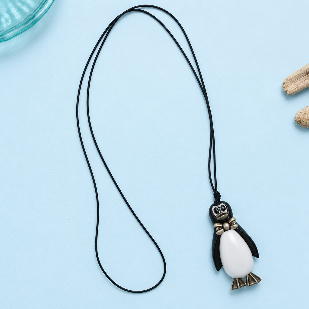 Pinguinkette mit lang ausgelegter schwarzer Kordel auf hellblauem Untergrund mit aquafarbenem Glas, Treibholz und hellen Steinen.