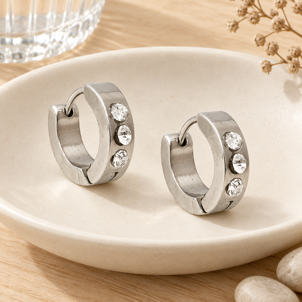 Dekobild: Silberfarbene Edelstahl-Creolen mit klaren Zirkonia-Steinen auf einer cremefarbenen Keramikschale mit Glas, Trockenblüten und hellen Steinen.