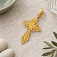 Filigraner Goldkreuz-Anhänger diagonal auf hellem Naturstein mit salbeifarbener Keramik, geriffeltem Glas, hellen Steinen und grünem Zweig.