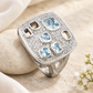 Silberring mit Aqua-Zirkonia an einem hellen Kiesel angelehnt, mit cremefarbenem Leinen und weißen Blüten